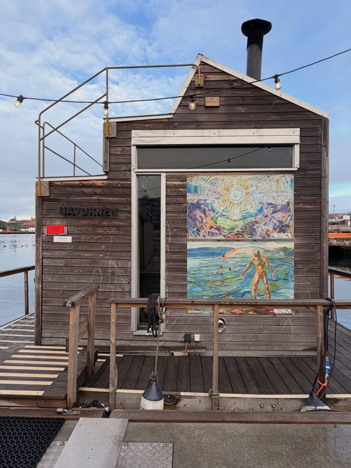 A wooden floating sauna with a painted mural, moored on the fjord