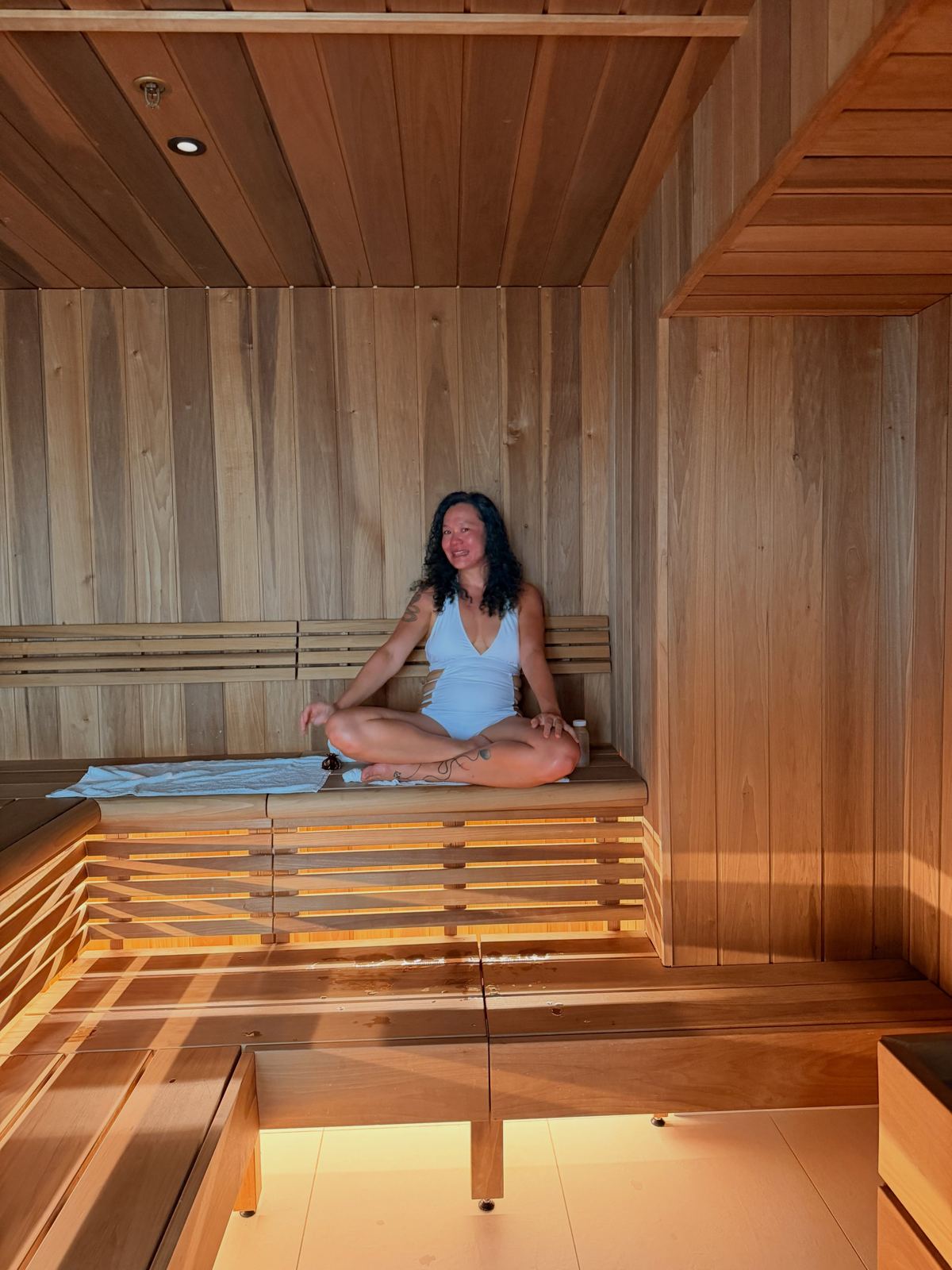 A quiet pale-wood sauna with a woman sitting cross-legged on the bench